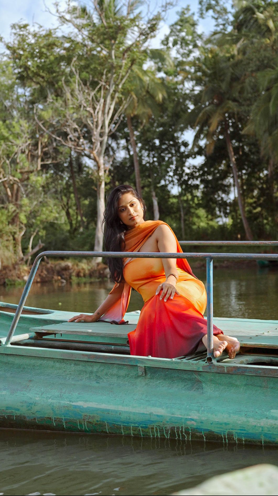 Model in an orange-to-red ombré drape seated on a turquoise boat on the river
