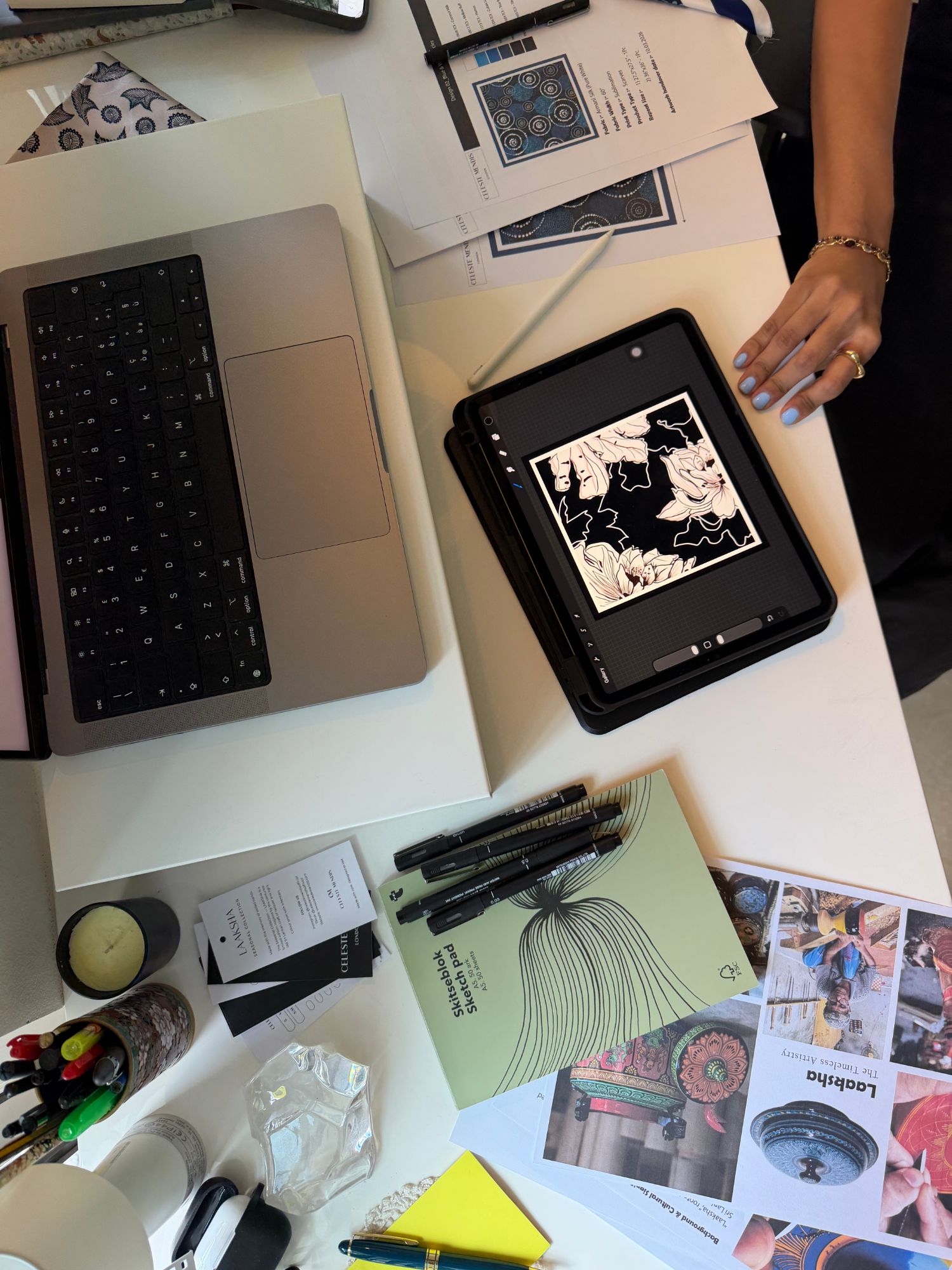 Studio desk with a floral print being drawn on a tablet, print tech-pack sheets and a sketch pad